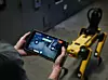 Zwei Hände halten ein Tablet, über das der Roboterhund Spot von Boston Dynamics im Hintergrund ferngesteuert wird
