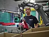 Ein Roboterarm hebt ein Plastikteil vom Fließband. Im Hintergrund ist eine Angestellte des Unternehmens zu erkennen.