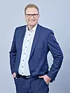 Mann mit Brille in blauem Anzug vor hellem Hintergrund, Hände in den Hosentaschen. Marco Grafe, Zeiss